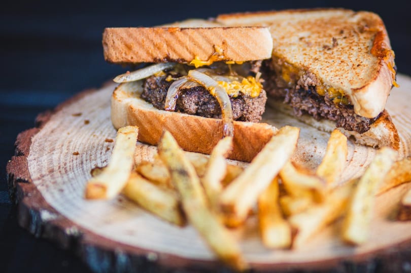 Patty melt with melted cheese and caramelized onions on toasted bread, served with fries on a wooden board.