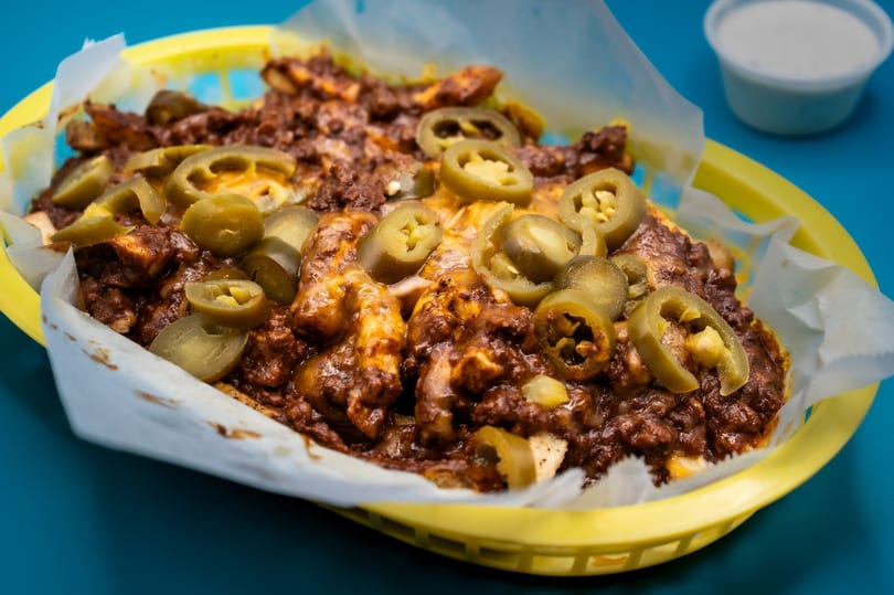 Chili cheese fries topped with sliced jalapeños in a yellow basket with a side of ranch.
