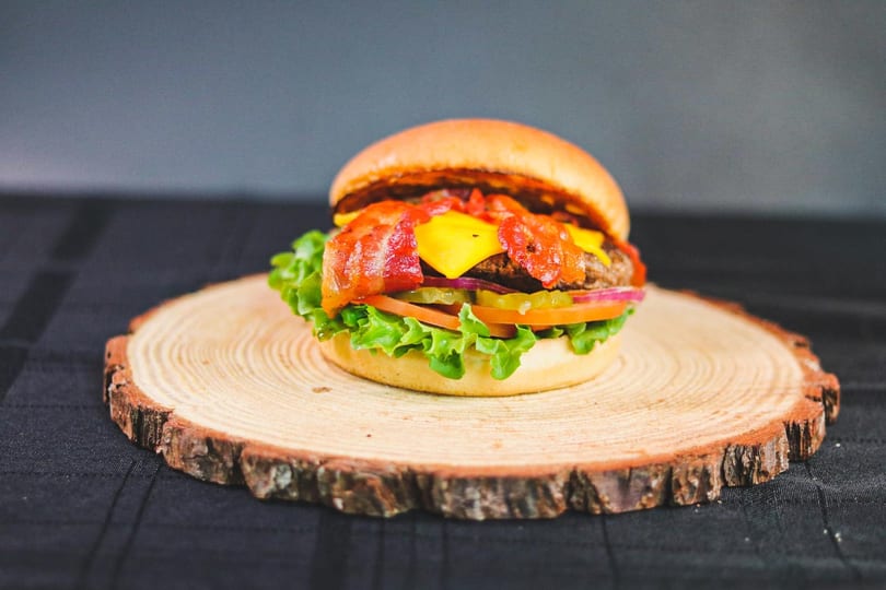 Bacon cheeseburger with lettuce, tomato, onion, and pickles on a wooden slab.